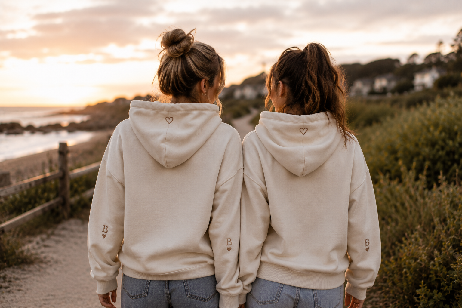 Zwei Freundinnen spazieren in farblich abgestimmten Hoodies gemeinsam auf einem Weg am Meer.